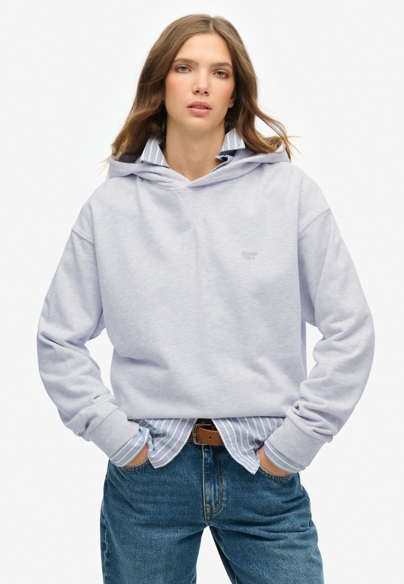 Sudadera gris claro hecha de un tejido suave con un bolsillo frontal, que cuenta con una capucha ajustable y un pequeño logotipo. Lleva puesta sobre una camisa a rayas azul.