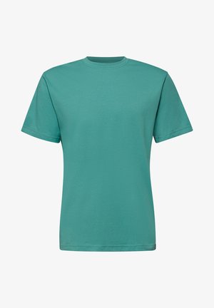 Junger Mann mit kurzen braunen Haaren, der ein türkisfarbenes kurzärmeliges Baumwoll-T-Shirt trägt und seitlich vor einem einfarbigen Hintergrund schaut.