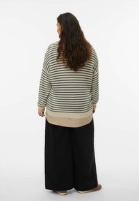Femme aux cheveux longs et ondulés portant un pull rayé crème et noir, une chemise beige en dessous, un pantalon large noir et des chaussures marron, debout devant un fond blanc.