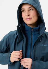 Giacca impermeabile blu navy con cappuccio, chiusura frontale con zip e polsini in velcro. Indossata sopra uno strato in pile blu con colletto alto.