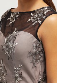 Šaty s průhledným černým výstřihem a květinovou výšivkou v odstínech stříbrné a bílé. Hladká šedá tkanina, přiléhavý střih, elegantní detaily.