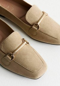 Mocassini in suede beige senza lacci con punta quadrata, caratterizzati da una barra in tonalità oro e dettagli cuciti lungo i bordi.