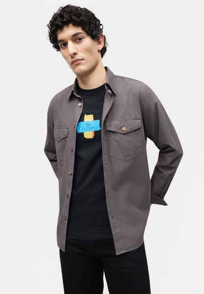 Νεαρός άνδρας με σγουρά μαλλιά φοράει γκρι πουκάμισο με κουμπιά ανοιχτό πάνω από μαύρο μπλουζάκι με σχέδιο, στέκεται μπροστά σε απλό λευκό φόντο.