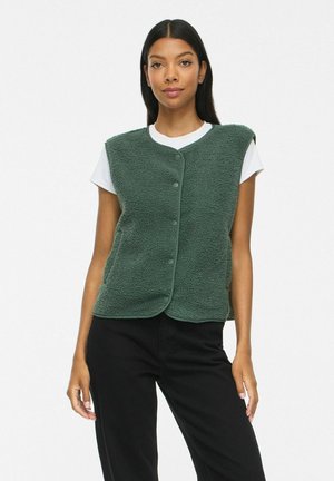 Vrouw draagt een donkergroen fleecevest met knopen over een wit shirt met korte mouwen en een zwarte broek, staand tegen een effen achtergrond.
