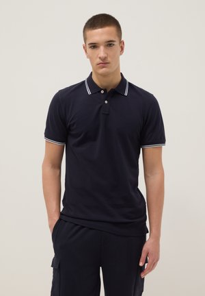 Junger Mann, der ein marineblaues Poloshirt mit weiß gestreiftem Kragen und Ärmeln sowie marineblaue Cargo-Hosen trägt, steht mit den Händen in den Taschen.