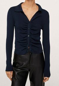 Camisa de botones de manga larga en color navy con detalles fruncidos en la parte delantera, textura de punto suave y cuello de camisa, combinada con pantalones ajustados negros.