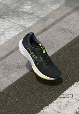 Schwarzer Laufschuh mit grünen und gelben Akzenten, Obermaterial aus Mesh, runde Zehenpartie und weiße Zwischensohle, auf Asphalt neben einer taktilen Gehfläche präsentiert.