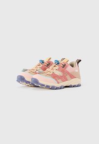 Paire de sneakers de trail en daim beige et rose avec semelles bleues antidérapantes et lacets élastiques sur fond blanc.