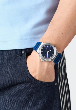 Uhr mit einem Gehäuse aus silbernem Edelstahl, einem blauen Gummireifen und einem dunkelblauen Zifferblatt, das silberne Stundenmarker und Zeiger aufweist.