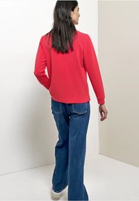 Rotes Sweatshirt mit langen Ärmeln, rundem Ausschnitt und geripptem Saum, kombiniert mit hoch taillierter, blauer Jeans mit weitem Bein und weißen Turnschuhen.