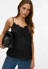 Czarna satynowa koszulka na cienkich ramiączkach z koronkowymi zdobieniami, gładka w dotyku, zestawiona z miękką czarną torebką typu worek, noszona do niebieskich jeansów.