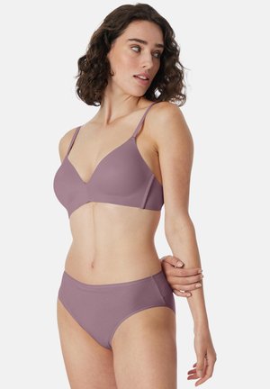 Woman with curly dark hair wearing mauve bra and matching underwear, standing with one arm crossed over her waist, looking sideways.