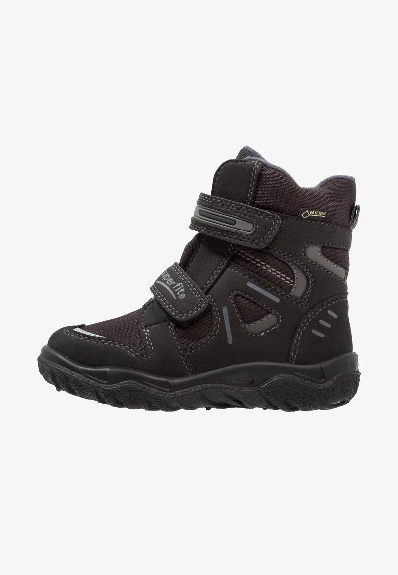 Bottine noire à bride en velcro. Fabriquée en daim et tissu, avec une semelle texturée, des surpiqûres contrastantes et une étiquette imperméable.