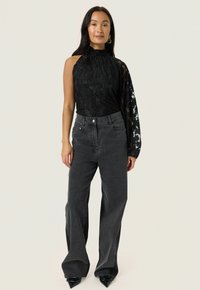 Schwarzes Spitzen-Haltertop mit durchsichtigen langen Ärmeln, kombiniert mit hoch taillierten grauen weiten Jeans und spitzen schwarzen Stiefeletten.