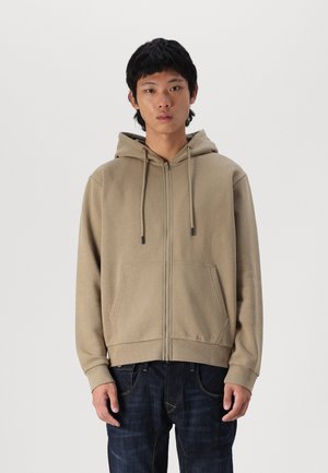 Beige Zip-Hoodie mit Kordelzugkapuze, Fronttaschen und gerippten Bündchen. Glatte Textur und lässiges Design. Getragen mit dunklen Jeans.