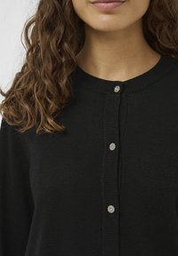 Fekete kardigán puha anyagból, kerek nyakkivágással és az elején öt virágmintás díszgombbal.