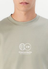 Nærbillede af en person iført en lysegrå T-shirt med rund hals, som har et hvidt logo med en globe, "CW", og teksten "Creative Works. Daily Essential Selections."
