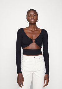 Top cropped preto de mangas longas com um recorte frontal e detalhe em anel metálico. Combinado com jeans brancos de cintura alta.
