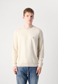 Ung mann iført en kremfarget langermet genser med en liten sirkulær logo på brystet og blå jeans, stående mot en ensfarget bakgrunn.