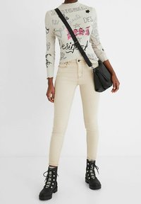 Haut gris clair à manches longues avec des motifs graphiques noirs et roses, associé à un jean skinny beige et des bottes noires à lacets. Sac bandoulière noir.