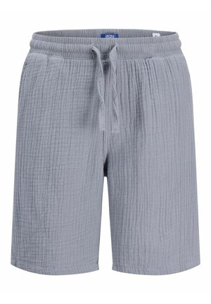 Chłopiec z kręconymi włosami stoi, ubrany w biały T-shirt z krótkim rękawem, szare krótkie spodenki, białe skarpetki i beżowe buty na jednolitym tle.