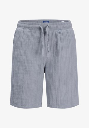 Chłopiec z kręconymi włosami stoi, ubrany w biały T-shirt z krótkim rękawem, szare krótkie spodenki, białe skarpetki i beżowe buty na jednolitym tle.