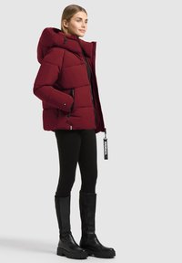 Femme portant une doudoune rouge foncé avec capuche, des leggings noirs et de grandes bottes noires, debout devant un fond clair uni.