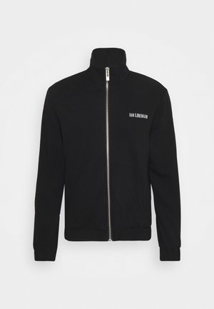 Mies, jolla on musta vetoketjullinen takki, valkoinen graafinen t-paita, siniset farkut ja mustat nahkasaappaat, seisoo valkoista taustaa vasten.