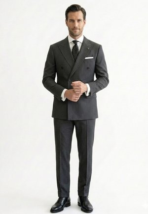 Hombre de pie con confianza, vestido con un traje cruzado de rayas finas gris oscuro, camisa blanca, corbata negra con lunares blancos y zapatos de vestir negros.