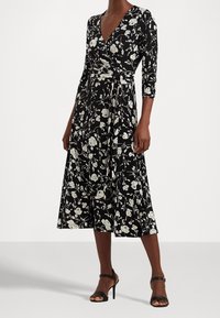 Czarna sukienka w kwiaty z rękawami o długości trzech czwartej, ozdobiona białymi wzorami róż oraz z wcięciem w talii, zestawiona z czarnymi sandałami na paski.