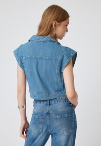 Skórzana kurtka z przyciętymi rękawami w jasnoniebieskim kolorze, z kołnierzem, szerokimi rękawami i surowym wykończeniem. Widoczna tekstura denimu, z detalami szwów z tyłu.