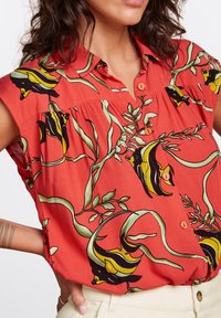 Camisa roja de manga corta con patrones de peces negros y amarillos y hojas verdes, con cierre de botones en la parte delantera y un ligero plisado debajo del cuello.