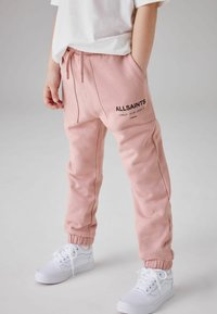 Růžové tepláky s elastickými manžetami a kapsami, se černým logem "ALLSAINTS". V kombinaci s bílými teniskami.