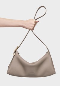 Mano sosteniendo un bolso de cuero texturizado color topo con una correa en forma de lazo sobre un fondo claro y liso.