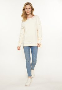 Felpa leggera color crema con dettagli in cucitura multicolore, maniche lunghe, spacchi laterali e vestibilità rilassata, abbinata a jeans skinny blu.