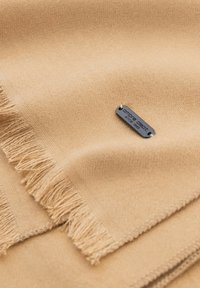 Beige uldhalstørklæde med frynser, der har et sort metalmærke med teksten "omb dept siden 1995" syet fast på stoffet.