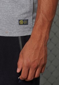 Camiseta gris con una etiqueta de tela que dice "ATHLETE SPORTS GOODS CO" y pantalones negros con un zipper plateado en el bolsillo lateral.