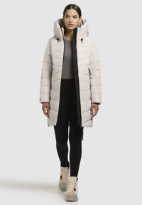Light beige puffer coat with a hood, vertical quilting, front zipper, and side pockets. Paired with black leggings and beige boots.