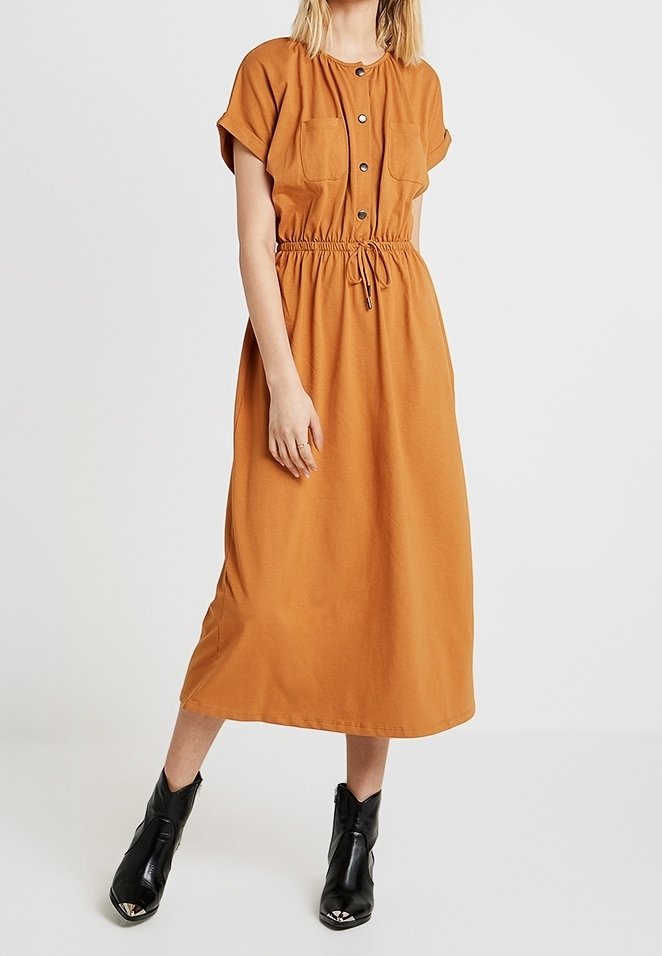 Robe midi couleur rouille avec manches courtes, boutonnée devant, deux poches poitrine et taille ajustable par cordon, portée avec des bottines noires.