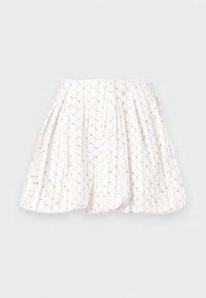 Žena stojaca s rukami na bokoch, oblečená v sivý pletený sveter, bielu vzorovanú sukňu a hnedé mokasíny na jednofarebnom pozadí.