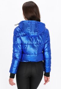 Giacca a vento blu con una finitura lucida e testurizzata, cappuccio e polsini elasticizzati. Design cropped con dettagli di cucitura orizzontale.