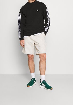 Černá mikina s bílým logem Adidas a třemi pruhy na rukávech, krémové šortky a zelené tenisky s bílými akcenty a hnědou podešví.