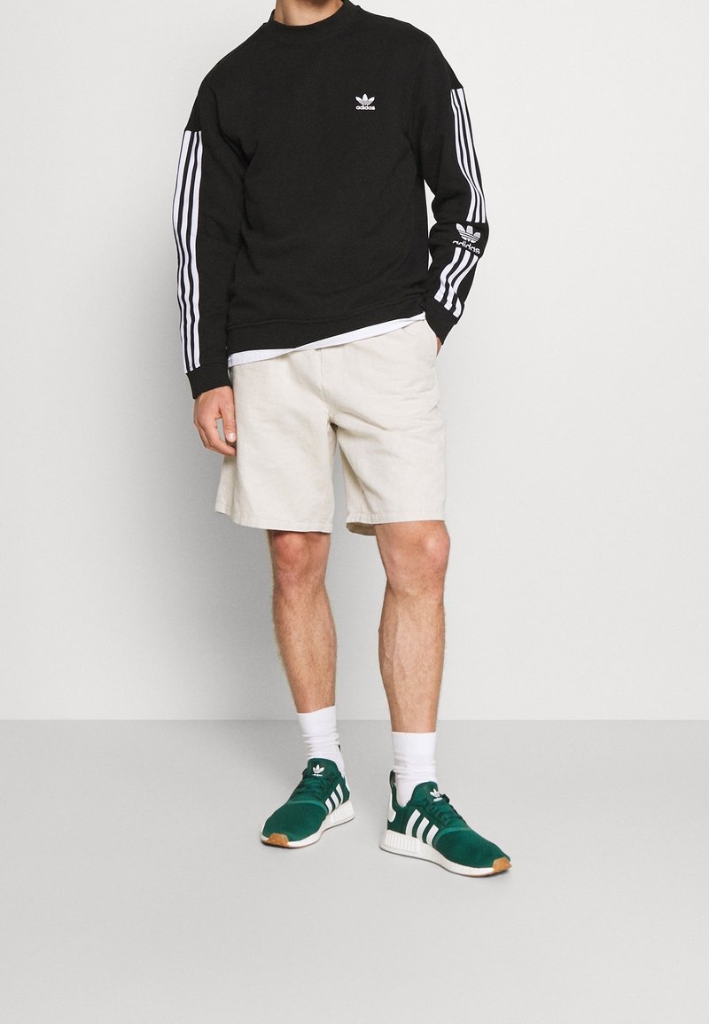 Felpa nera con logo Adidas bianco e maniche a tre strisce, pantaloni corti crema e sneakers verdi con dettagli bianchi e suola in gomma.