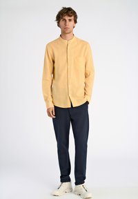 Camicia button-up di colore giallo chiaro con colletto alla cinese, tasca sul petto; abbinata a pantaloni blu navy e sneakers bianche.