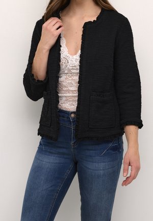 Mujer con chaqueta negra texturizada con bolsillos sobre una camiseta blanca de encaje y vaqueros azules, de pie frente a un fondo claro y liso.
