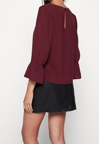 Burgundin paita, jossa on kellotyttöhiha, teksturoitu kangas ja avattava selkäkohta, yhdistetty mustaan nahkahameeseen.