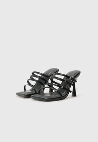 Mules à talons noirs avec plusieurs fines lanières et un design à bouts carrés. Texture lisse avec des accents de marque discrets sur les lanières.