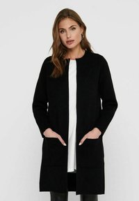 Black long cardigan with a structured design, open front, two front pockets, and a smooth texture. Worn over a white shirt and black pants.