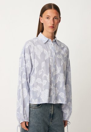 Jeune femme aux cheveux bruns raides portant une chemise ample à boutons bleu et blanc à motif floral et un jean bleu, debout devant un fond uni.