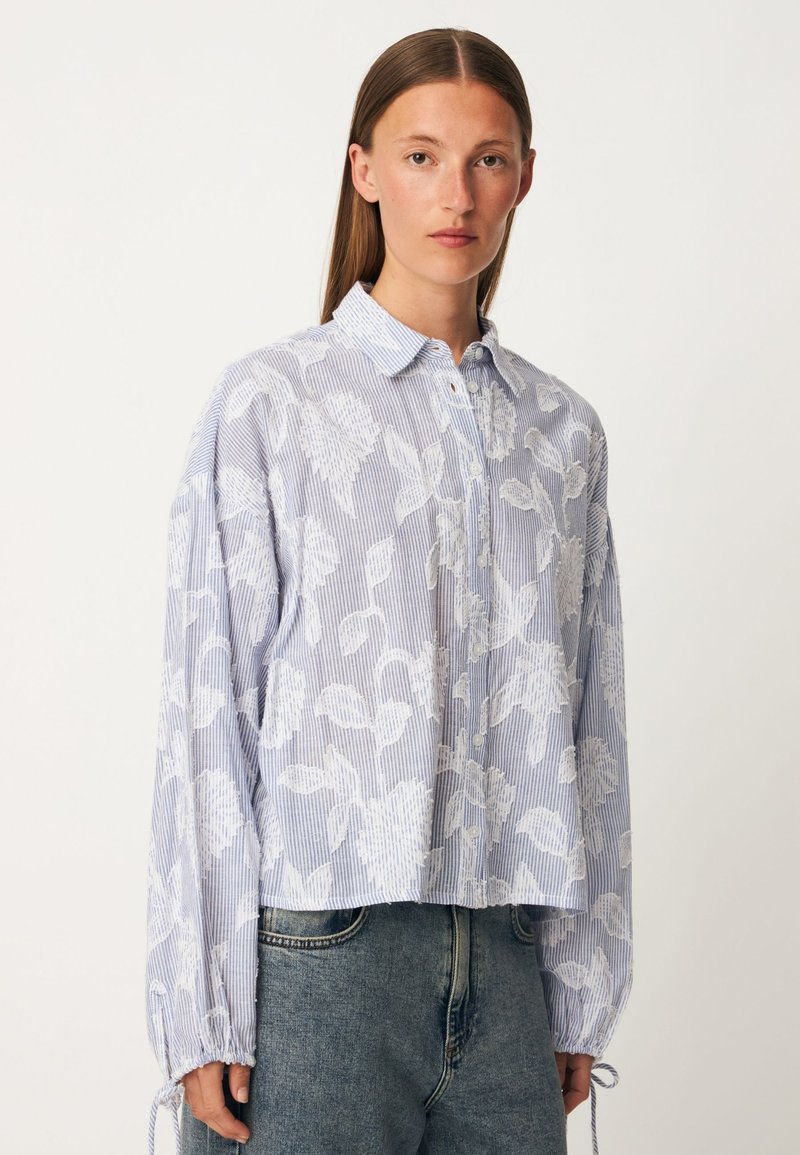 Jeune femme aux cheveux bruns raides portant une chemise ample à boutons bleu et blanc à motif floral et un jean bleu, debout devant un fond uni.